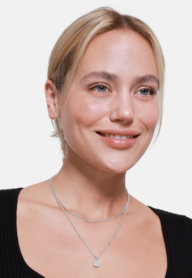 Zilveren gelaagde necklaces met een getextureerde hanger aan de onderste ketting, gedragen over een zwart geribd topje. Glad metalen oppervlak en slank design.