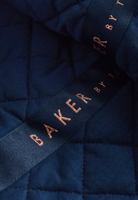 Ciemnoniebieska pikowana tkanina z czarną wstążką ozdobioną miedzianym napisem Baker by Ted Baker.