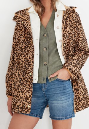 Une femme porte une veste imprimée léopard par-dessus une chemise blanche à col et un haut vert boutonné avec un short en jean bleu, main dans la poche.