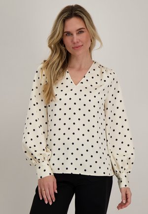 Vrouw met lang blond haar die een witte blouse met zwarte stippen en een zwarte broek draagt, staand tegen een effen achtergrond.