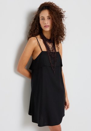 Donna con capelli ricci che indossa un vestito nero senza maniche e una collana nera a strati con perline, in piedi contro un muro bianco.