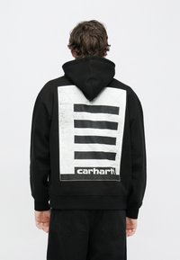 Sudadera negra con capucha con un gran gráfico blanco que presenta franjas horizontales negras y el logo "carhartt" en la espalda. Material de algodón.