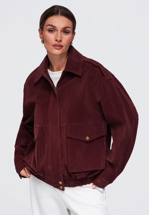 Donna con capelli castani che indossa una giacca oversize color bordeaux con grandi tasche frontali e pantaloni bianchi su uno sfondo chiaro e uniforme.