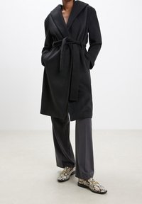 Osoba oblečená v černém kabátu s páskem, tmavých širokých kalhotách a mokasínech s hadím potiskem a zlatými řetízkovými detaily, stojící před jednobarevným pozadím.