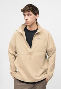 Νέος άνδρας με κοντά σγουρά μαλλιά φοράει μπεζ τζάκετ με φερμουάρ στο μισό πάνω μέρος πάνω από μαύρο πουκάμισο, τα χέρια στις τσέπες, κοιτάζοντας αριστερά μπροστά σε απλό φόντο.