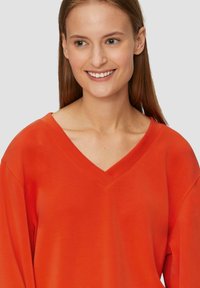 Bluza z dekoltem w serek w intensywnym pomarańczowym kolorze, wykonana z miękkiego materiału. Posiada długie rękawy i luźny krój.