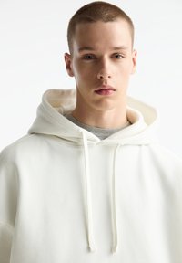 Hanorac alb cu glugă din material moale, cu un croi lejer, șnururi și un guler gri stratificat. Design simplu, minimalist.