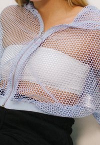 Zbliżenie na osobę w jasnoniebieskiej siateczkowej bluzie na zamek, założonej na biały biustonosz bez ramiączek i czarne spodnie.