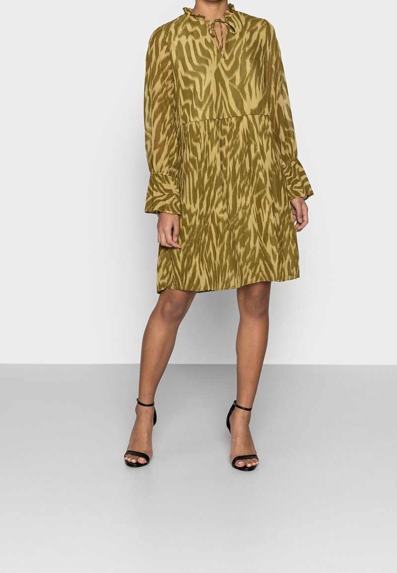 Femme portant une robe longue jusqu'aux genoux à manches longues vert olive à motifs avec des poignets à volants et des talons noirs à bride cheville, debout sur un sol gris.