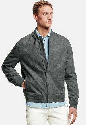 Jonge man draagt een grijze jas met rits over een lichtblauw overhemd en witte broek, met één hand in zijn zak, kijkt naar rechts.