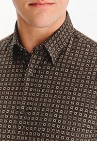 Zbliżenie dolnej części twarzy i szyi mężczyzny w zapinanej ciemnej koszuli z małym geometrycznym wzorem w kształcie gwiazdy.