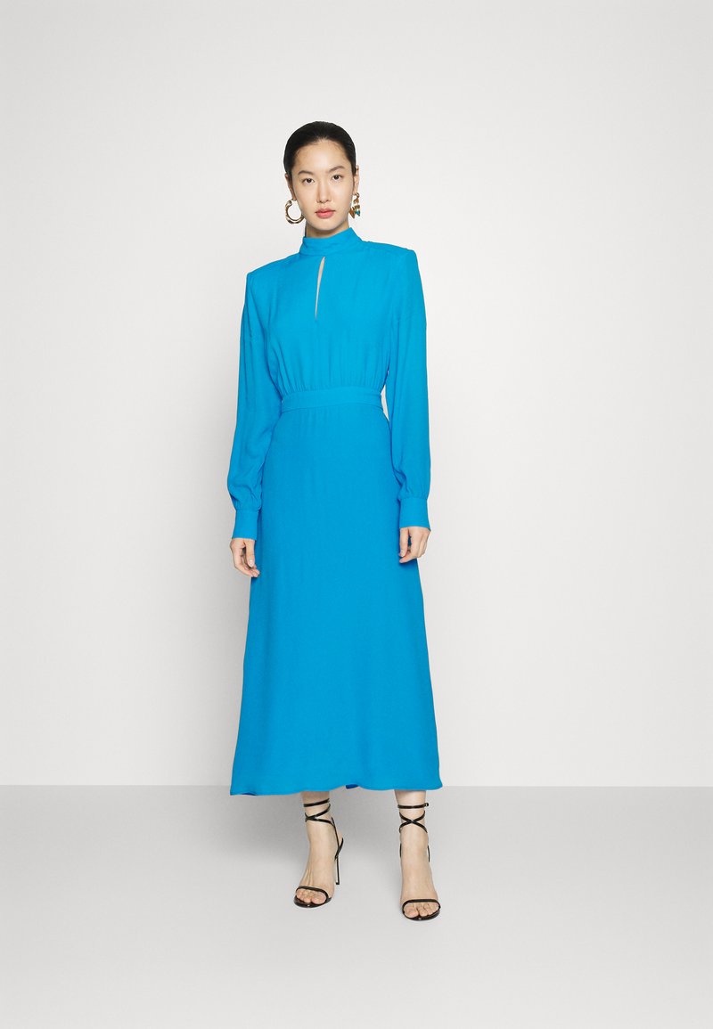 Niebieska sukienka z długimi rękawami, wysokim kołnierzem, detalem w kształcie otworu, dopasowaną talią i zwiewną spódnicą; zestawiona z czarnymi sandałami na obcasie z paskami.