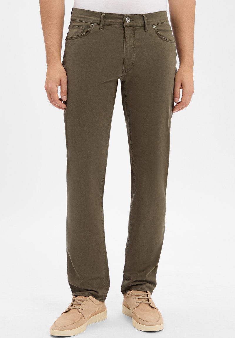 Uomo che indossa pantaloni aderenti verde oliva e scarpe beige stringate, in piedi contro uno sfondo bianco.