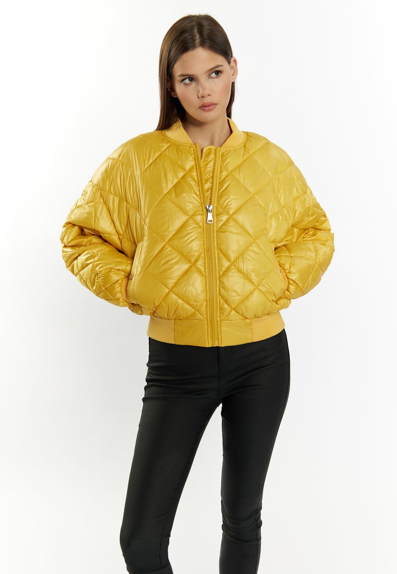 Giacca bomber trapuntata gialla con zip frontale, polsini e orlo a costine, caratterizzata da una texture lucida e un motivo a diamante. I pantaloni neri aderenti completano il look.