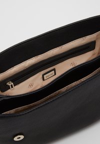 Interno di una borsa a tracolla in finta pelle nera con una fodera beige, dotato di una tasca con zip e un'etichetta metallica di marca.
