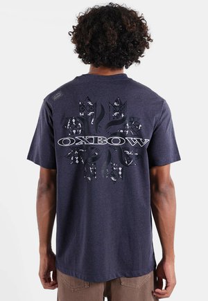 Adulte aux cheveux bouclés portant un t-shirt bleu foncé Oxbow avec un grand logo à motifs dans le dos, se tenant devant un fond uni.