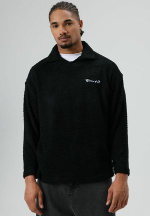 Zwarte fleece trui met een losse pasvorm, voorzien van een kraag, lange mouwen en witte borduursels op de borst. Zachte textuur, minimalistisch ontwerp.