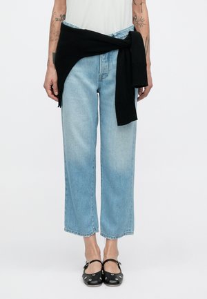 Lyseblå jeans med vide ben og en falmet finish, kombineret med en sort sweater bundet ved taljen. Sorte flade sko med dekorative hardware-detaljer.