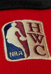 Roter Stoff mit einem gestickten NBA-Logo in Blau, Kastanienbraun und Gold. Das Logo zeigt die Silhouette eines Spielers und den Text "HWC".