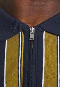 Marinblå zip-up polo med gröna och gula vertikala ränder. Har en stickad textur, krage och detaljer med metallisk dragkedja.