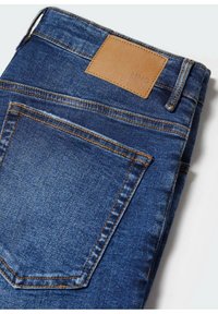 Skládané modré džíny z denimu s viditelnou zadní kapsou, poutkem na opasek a prošitým hnědým koženým znakem na pase.