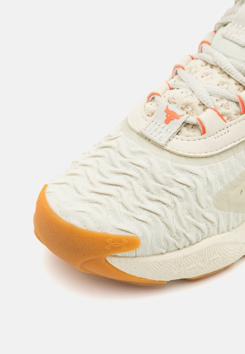 Jasne beżowe buty sportowe z teksturowaną cholewką, pomarańczową podeszwą i wyraźnym detalem logo. Charakteryzują się plecionym wzorem i kontrastującymi sznurówkami.