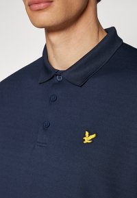 Námornícky polo tričko vyrobené z texturovanej látky, s trojitém gombíkovom zapínaní a žltým logom orla na hrudi.