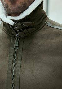 Chaqueta verde oliva con un alto collar forrado de piel sintética blanca, cremallera de metal y detalles laterales. Textura suave con un acabado mate.