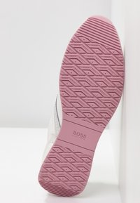 Semelle de basket blanche avec une semelle rose présentant des rainures diagonales et un motif de texte répété « HUGO BOSS », logo de la marque au centre.