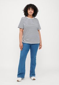 T-shirt rayé bleu marine et blanc, manches courtes, col rond. Associé à un jean bleu évasé et des baskets blanches. Cheveux bouclés encadrant le visage.