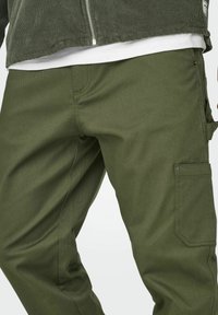Pantalones cargo color verde oliva con una textura suave, que cuentan con bolsillos laterales y un brillo sutil. Combinados con una chaqueta con cremallera en capas.