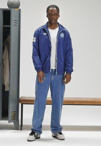 Marineblå vindbreaker med hvite detaljer, glidelås foran og logo på brystet; kombinert med lyseblå jeans og svarte joggesko. Ensfarget bakgrunn.