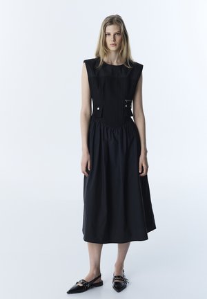 Mujer joven de pie con un vestido midi negro sin mangas, hombros estructurados y bolsillos delanteros, llevando zapatos planos negros con punta y hebillas.