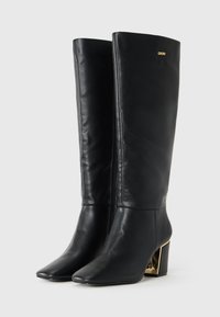 Stivali di pelle neri al ginocchio con punte appuntite, tacco a blocco con accenti metallici dorati e un piccolo logo DKNY vicino alla parte superiore.