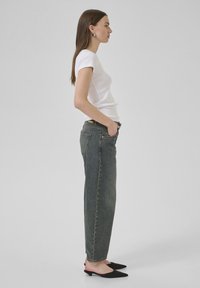 Baltas īsām piedurknēm T-krekls, kas apvienots ar augsto jostasvietu, izbalējušām zilām taisnām biksēm. Modelis valkā melnus asus apavus un stāv profilā.