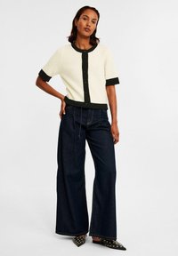Kvinna som bär en kortärmad krämfärgad cardigan med svarta detaljer, vida mörk denimjeans och svarta spetsiga flats med nitar, stående med handen på höften.