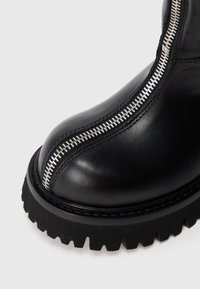 Schwarzer Lederstiefel mit dickem Profilsohlen und zentralem silbernem Reißverschluss, der von der Zehe bis zum Knöchel verläuft.