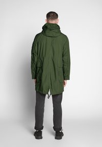 Grön parka-jacka med huva, med insydd midja, sidofickor och längre bak. Tyget ser slätt och lätt ut.