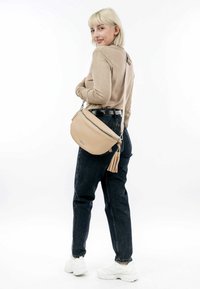 Beige leren schoudertas met een franjes, met een gladde textuur en een gebogen ontwerp, gecombineerd met donkere jeans en witte sneakers.