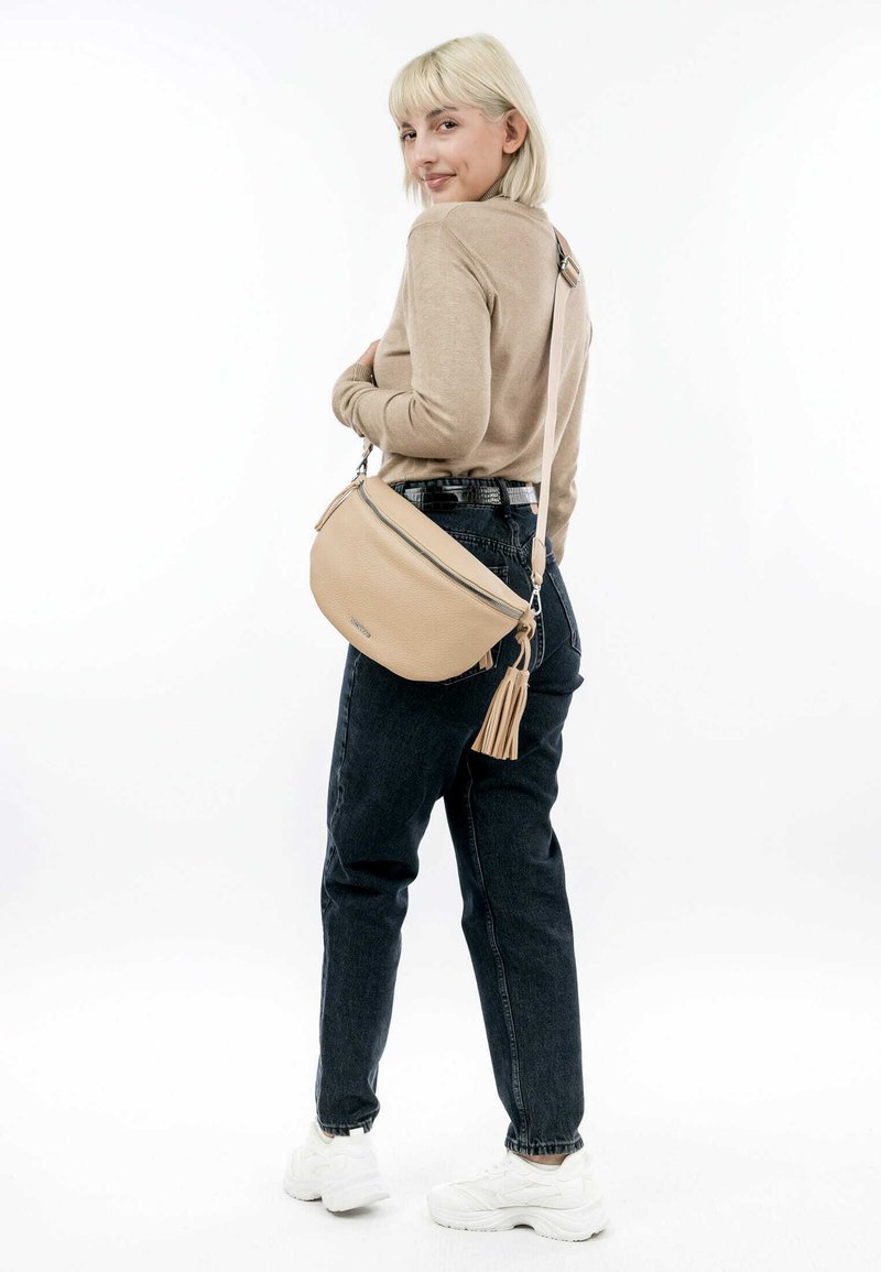 Beige leren schoudertas met een franjes, met een gladde textuur en een gebogen ontwerp, gecombineerd met donkere jeans en witte sneakers.