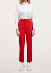 Pantalones rojos de corte ajustado con cintura alta, pliegues delanteros y cierre de un solo botón, combinados con una camisa blanca de cuello y zapatillas blancas.