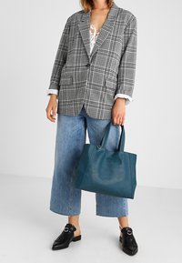 Šedé kostkované sako s vykrojenými klopami, v kombinaci se širokými modrými džínami. V ruce drží petrolejovou koženou kabelku s hladkou texturou a dvojitými uchy.