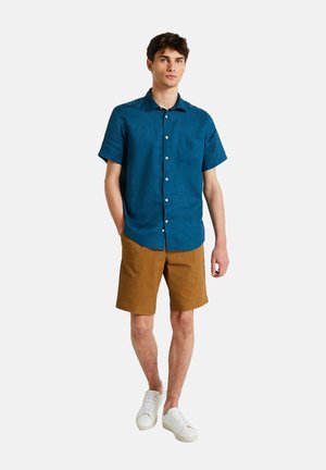 Chemise bleue à manches courtes avec un devant à boutons et une poche, associée à un short marron et des baskets blanches. Texture de tissu lisse, design décontracté.
