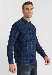 Chemise en flanelle à carreaux bleu marine avec deux poches poitrine, col boutonné et manches longues. Présente des motifs à carreaux bleu clair et gris.