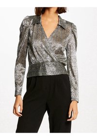 Frau in einer langärmligen, silbernen Metallic-Wickelbluse und schwarzen Hosen, stehend vor einem schlichten weißen Hintergrund.