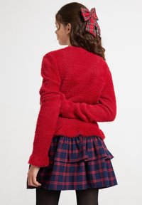 Roter, flauschiger Pullover mit langen Ärmeln und geschichtetem Saum, kombiniert mit einem marineblauen Karottenschottenrock mit roten Akzenten und Rüschen. Haare geschmückt mit einer karierter Schleife.