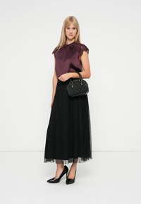 Blusa di raso bordeaux con bordo in pizzo, abbinata a una gonna maxi in tulle nera e tacchi alti neri; tiene in mano una borsa nera strutturata.