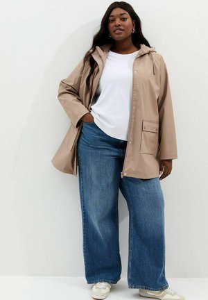 Vrouw die een beige regenjas met capuchon draagt, een wit T-shirt, wijde blauwe jeans en witte sneakers, staand tegen een witte muur.