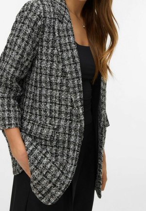 Frau trägt einen schwarz-weiß strukturierten Blazer über einem schwarzen Top, mit einer Hand in der Blazertasche, lange braune Haare sichtbar.
