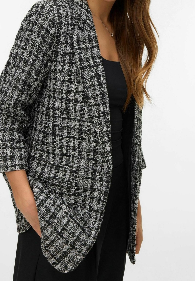 Femme portant un blazer texturé noir et blanc sur un haut noir, une main dans la poche du blazer, cheveux longs bruns visibles.